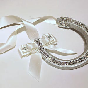 May include: A silver horseshoe adorned with sparkling rhinestones and a white satin ribbon. The horseshoe features a bow embellished with rhinestones. The ribbon is looped and tied in a bow. A symbol of good luck.