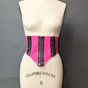 Peut inclure: Un corset rose et noir avec des œillets et des lacets noirs. Le corset est sur un mannequin blanc.