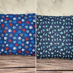 Op de afbeelding: Twee blauwe kussenhoezen met verschillende patronen. Eén heeft een pootprintpatroon in rood, blauw, wit en grijs. De andere heeft een bot- en pootprintpatroon in blauw, wit en grijs.