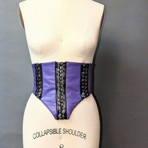 Peut inclure: Corset en cuir violet avec laçage noir et œillets argentés. Le corset est sur un mannequin blanc.