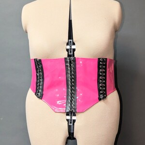 Peut inclure: Un corset en cuir verni rose avec des détails en dentelle noire et des œillets argentés.