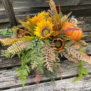 May include: A fall-themed floral arrangement featuring sunflowers, pumpkins, and various types of foliage. The arrangement includes bright yellow sunflowers, an orange pumpkin, and green and brown leaves. The arrangement is set on a weathered wooden surface.