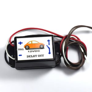 May include: A black electronic device with a white label featuring an orange car graphic and the text "12VDC DELAY OFF". The device has red, white, and brown wires attached, and is likely an automotive component.