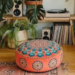 Pode incluir: Uma almofada de chão redonda com uma base de tecido laranja vibrante e um padrão de mandala detalhado e simétrico em tons de azul, turquesa e creme. As bordas são finalizadas com um acabamento decorativo de pequenos pompons turquesa.