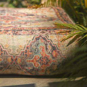 May include: Close-up of a decorative, rectangular cushion with a detailed, colorful pattern. The design features a mix of red, blue, yellow, and black elements on a beige background. The cushion is partially obscured by green foliage.