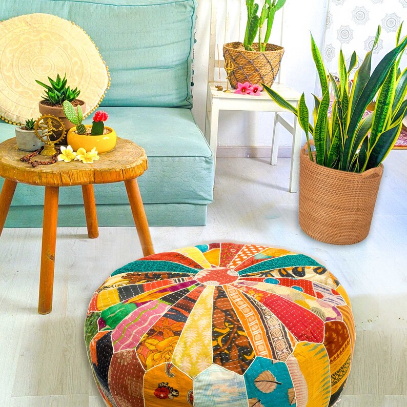 May include: A colorful patchwork floor cushion with a variety of patterns and colors, including red, yellow, blue, and green. The cushion is sitting on a light wood floor in a room with plants and furniture.