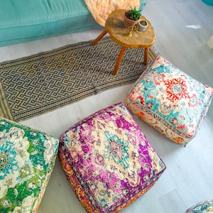 May include: Overhead shot of a living room setup featuring four square floor cushions with intricate, colorful patterns. A patterned rug and a small wooden side table with a plant and decor are also visible. The cushions have a vintage, bohemian aesthetic.