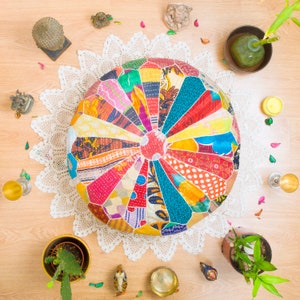 May include: A colorful, round, patchwork floor cushion with a white crocheted doily underneath. The cushion is made of various fabrics in shades of red, blue, green, yellow, and orange. The cushion is surrounded by decorative items, including a small potted plant, a candle, and a few other decorative objects.