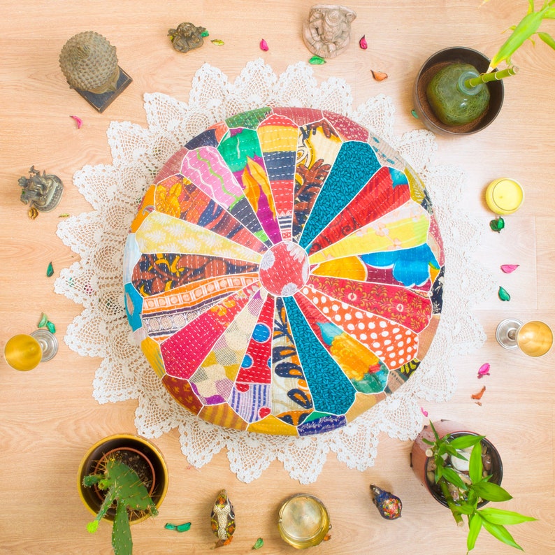 May include: A colorful, round, patchwork floor cushion with a white crocheted doily underneath. The cushion is made of various fabrics in shades of red, blue, green, yellow, and orange. The cushion is surrounded by decorative items, including a small potted plant, a candle, and a few other decorative objects.