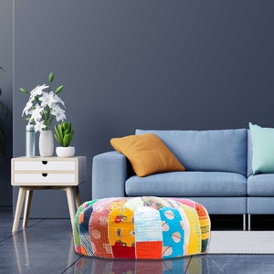 May include: A colorful patchwork round pouf ottoman with a blue, yellow, red, and green pattern. The pouf is sitting in front of a blue sofa with a yellow pillow.