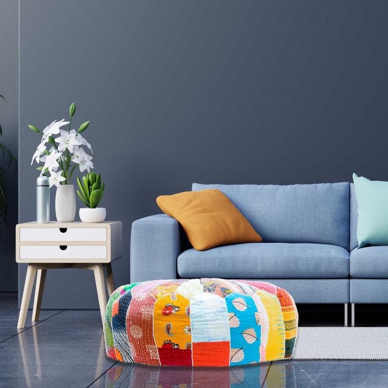 May include: A colorful patchwork round pouf ottoman with a blue, yellow, red, and green pattern. The pouf is sitting in front of a blue sofa with a yellow pillow.