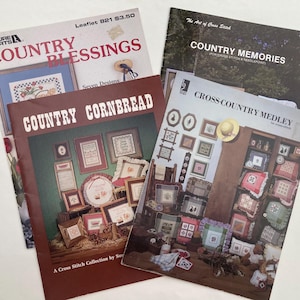 May include: A collection of vintage cross-stitch pattern booklets. Titles include "Country Blessings," "Country Cornbread," "Country Memories," and "Cross-Country Medley." The covers feature images of framed cross-stitch samplers and country-themed designs.