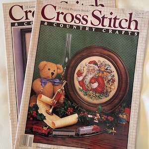 May include: Three vintage "Cross Stitch & Country Crafts" magazines. The covers feature a cross-stitch design of Santa Claus, a teddy bear, and miniature train set. The magazines are from the 1980s and have a grid pattern.