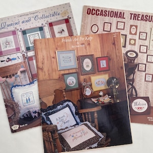 May include: Three vintage craft books are displayed, featuring cross-stitch patterns. The covers showcase framed samplers, pillows, and decorative items. Titles include "Quaint and Collectible" and "Occasional Treasures."