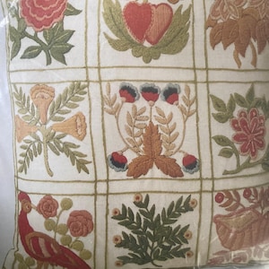 Könnte beinhalten: Quadratisches Zierkissen mit gestickten Blumen- und Vogelmotiven. Das Kissen hat ein Rastermuster mit verschiedenen Stickmotiven, darunter Rosen, Herzen und ein Vogel. Die Farbpalette umfasst Rot, Grün, Orange und Creme.