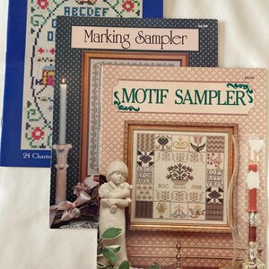 May include: Three vintage cross-stitch pattern books. The blue book reads "Easy-to-Make Cross-Stitch Samplers and Mottoes." The other two books are titled "Marking Sampler" and "Motif Sampler's." A candle and a statue are also in the image.