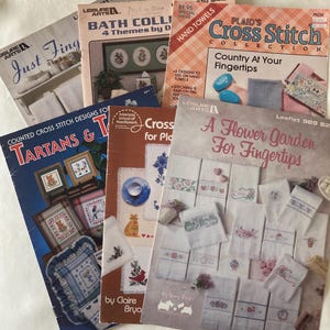 May include: A collection of vintage cross-stitch pattern books. Titles include "Bath Collection," "Cross Stitch Collection," and "A Flower Garden for Fingertips." The covers feature images of finished cross-stitch projects, including floral and tartan designs.