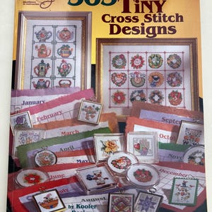 May include: A book titled "365 Tiny Cross Stitch Designs" with framed cross-stitch samplers and small, framed cross-stitch pendants. The book cover is yellow and features cross-stitch designs of teapots and floral wreaths. Pendants are displayed with calendar months.