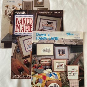 May include: A collection of vintage cross-stitch pattern books with country themes. Titles include "Along the Countryside," "Baked in a Pie," and "Down a Farm Lane." The covers feature framed needlework and rustic imagery.