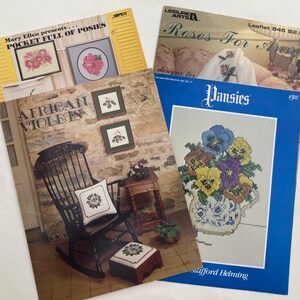 May include: A collection of vintage cross-stitch pattern booklets. Titles include "Pocket Full of Posies", "African Violets", "Roses For Anne", and "Pansies". The booklets feature floral designs and illustrations, with a rustic aesthetic.
