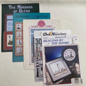 May include: A collection of cross-stitch pattern books featuring coastal themes. Titles include "The Marshes of Glynn," "Lighthouses of the Great Lakes," and "Beacons by the Shore." The covers display lighthouse imagery and nautical designs.