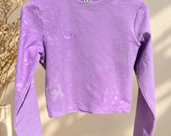 May include: A long-sleeve crew neck shirt in a vibrant purple hue, featuring a subtle, mottled bleach-wash effect across the fabric. The garment is displayed on a wooden hanger, showcasing a cropped hemline and a simple, clean neckline.