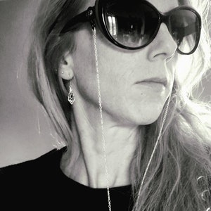 Peut inclure: Une femme aux longs cheveux blonds portant des lunettes de soleil noires avec une chaîne argentée attachée aux montures. Elle porte un chemisier noir.