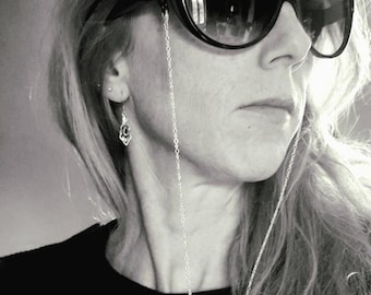 May include: A woman with long blonde hair wearing black sunglasses with a silver chain attached to the frames. She is wearing a black shirt.