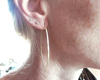 May include: A long, thin, gold-colored metal earring with a curved, hammered texture. The earring is shaped like an elongated crescent or arch, hanging vertically from the earlobe. The surface of the metal has a subtle, uneven, reflective finish.