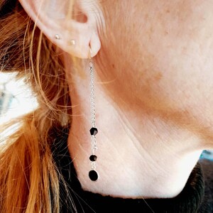 Peut inclure: Boucle d'oreille longue en chaîne argentée ornée de perles noires et d'un pendentif ovale noir. La boucle d'oreille est suspendue à une oreille percée. La personne porte un haut noir.