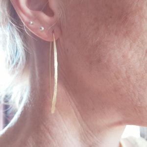 May include: A large, thin, circular hoop earring with a textured, hammered gold-tone finish. The earring is suspended from the earlobe, featuring a simple wire construction that maintains a round shape and a reflective, metallic surface.