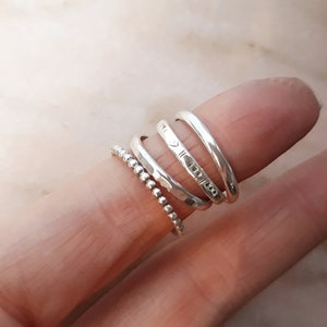 May include: A selection of silver rings on a finger. The rings include a beaded band, a hammered band, and several smooth bands with etched designs. The rings are made of a shiny, metallic material.