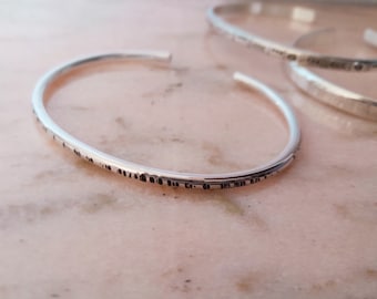 May include: A set of three silver-toned open cuff bracelets. The front bracelet has a textured pattern along the bottom edge. The bracelets are displayed on a pink and white marble surface. Jewelry.