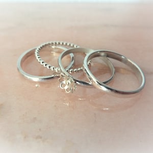 May include: Four silver rings of different designs are displayed on a pink surface. One ring has a beaded texture, another has a floral design, and the other two are plain bands. The rings are likely made of sterling silver.