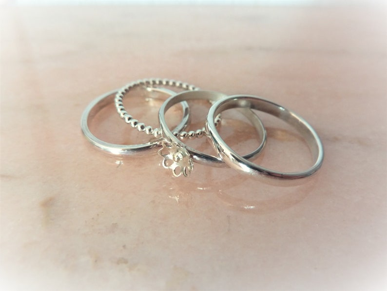 May include: Four silver rings of different designs are displayed on a pink surface. One ring has a beaded texture, another has a floral design, and the other two are plain bands. The rings are likely made of sterling silver.