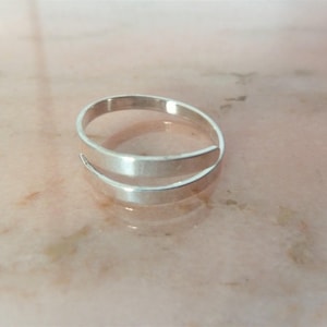 Pode incluir: Um anel de prata com um design simples e minimalista. O anel é feito de duas bandas que envolvem o dedo.