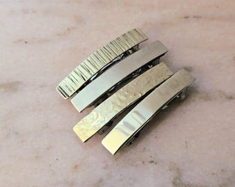 May include: A set of four rectangular metal hair barrettes with varying surface textures. The clips feature finishes including vertical lines, a smooth matte surface, a hammered effect, and a polished reflective gold-tone surface.
