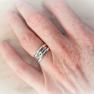 May include: Three silver rings, one with a small, pale floral design. The rings are stacked on a finger, showing the intricate details of the jewellery. The image is well-lit, highlighting the rings' shine and the floral accent.