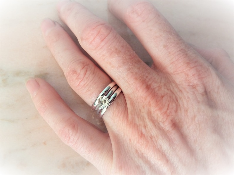 May include: Three silver rings, one with a small, pale floral design. The rings are stacked on a finger, showing the intricate details of the jewellery. The image is well-lit, highlighting the rings' shine and the floral accent.