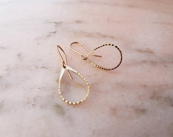 May include: A pair of gold-toned teardrop-shaped earrings featuring a textured, hammered surface. Each earring is suspended from a curved wire hook, creating an open, minimalist loop design that hangs freely from the top attachment point.