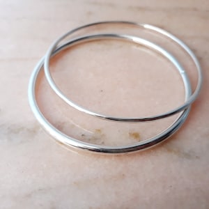May include: Two silver bangle bracelets, one slightly larger than the other, are stacked on top of each other. The bracelets are smooth and shiny.