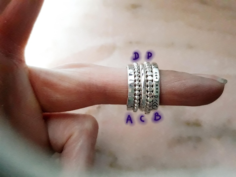 May include: A selection of silver rings displayed on a finger. The rings showcase diverse designs, such as hammered textures, beaded details, and dotted patterns. The rings are set against a soft, blurred background.