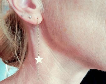 May include: A gold-colored earring with a star pendant hangs from a person's ear. The earring features a delicate chain and a small star charm. Two small silver studs are also visible in the ear. The background is a soft, neutral tone.