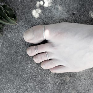 Peut inclure: Image en niveaux de gris d'un pied orné de trois bagues d'orteil argentées. Les bagues sont fines et simples, une sur le gros orteil et deux sur l'orteil adjacent. Le pied repose sur une surface grise texturée.