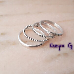 May include: A set of silver rings with different patterns. One ring has small beads, another has an arrow design, and the others are plain bands. The rings are stacked on a light pink surface. The text "Compo G" is visible.