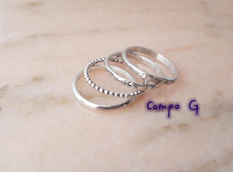 May include: A set of silver rings with different patterns. One ring has small beads, another has an arrow design, and the others are plain bands. The rings are stacked on a light pink surface. The text "Compo G" is visible.