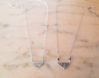 May include: Two silver necklaces with triangular pendants. One pendant has a textured, cracked pattern, while the other has a linear texture and small black beads. Both necklaces are displayed on a light-colored surface.