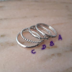 May include: Four silver rings are shown on a light pink surface. The rings feature various designs: a plain band, a beaded band, a band with a chevron pattern, and a thin band. The rings are labelled A, B, C, and D.