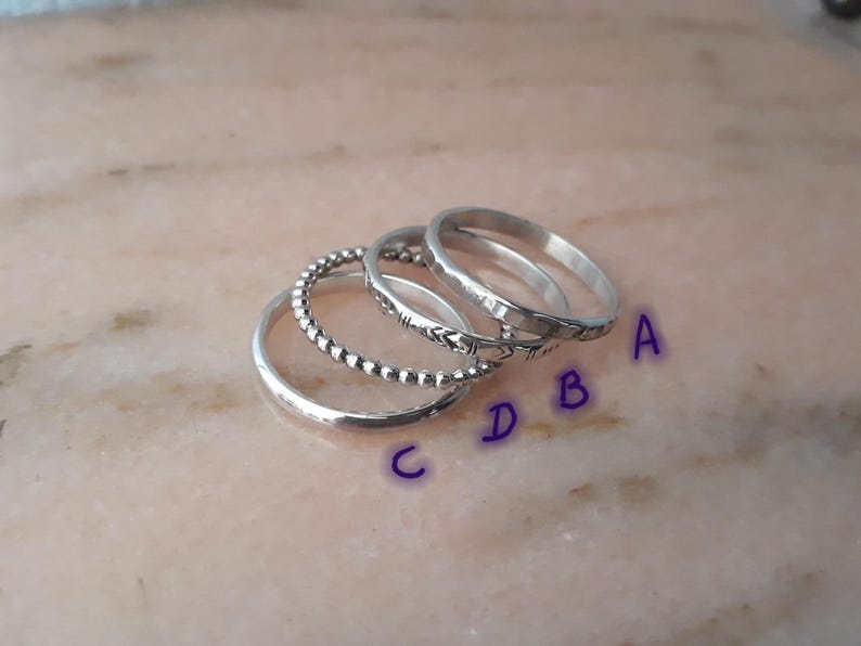 May include: Four silver rings are shown on a light pink surface. The rings feature various designs: a plain band, a beaded band, a band with a chevron pattern, and a thin band. The rings are labelled A, B, C, and D.