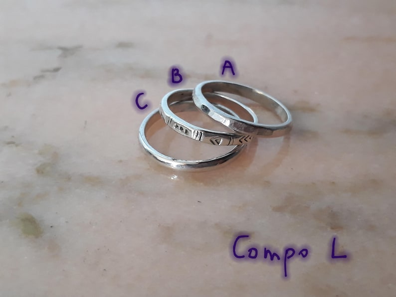 May include: Three silver rings are shown, each with a distinct design. Ring A has a hammered texture, Ring B features geometric etchings, and Ring C has a smooth, polished surface. The rings are arranged on a light-coloured surface. The text "Compo L" is written in purple.